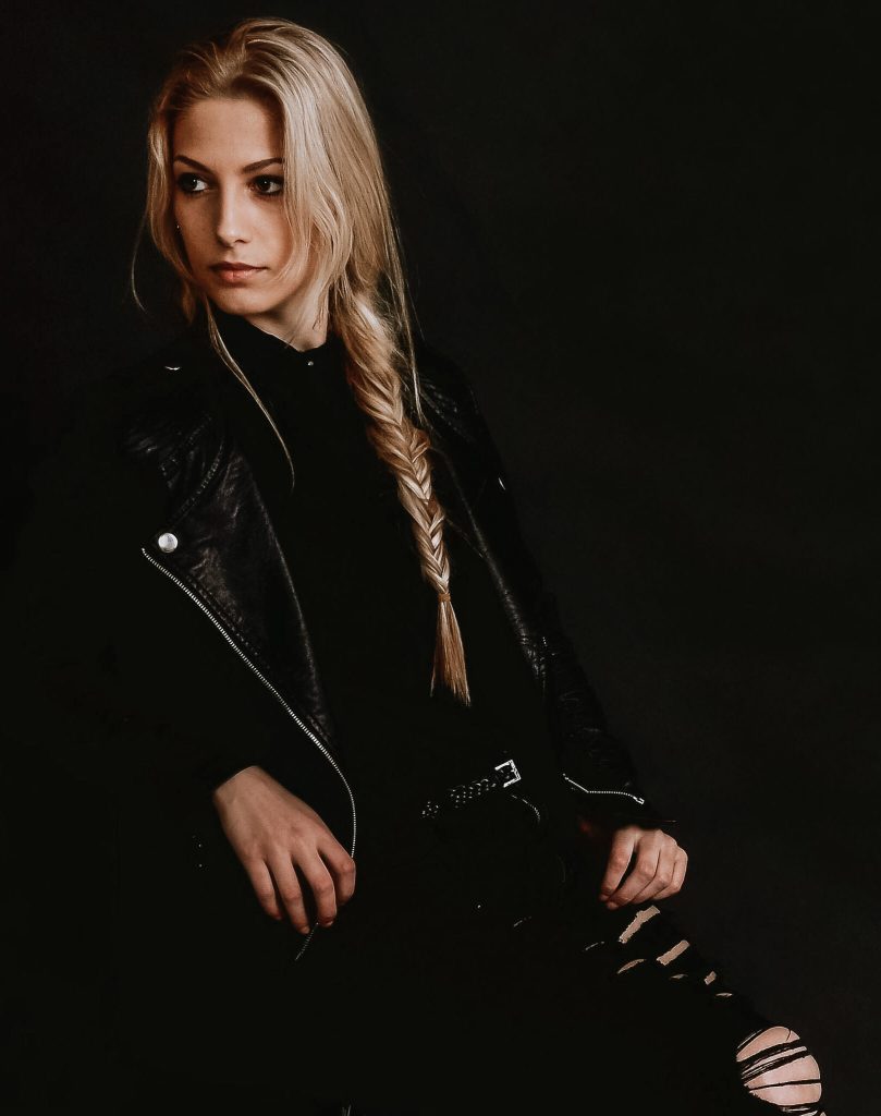 Profile portrait of Laurine, wearing a black leather moto-style jacket and shredded jeans, captured against a deep, dark studio backdrop with moody lighting.