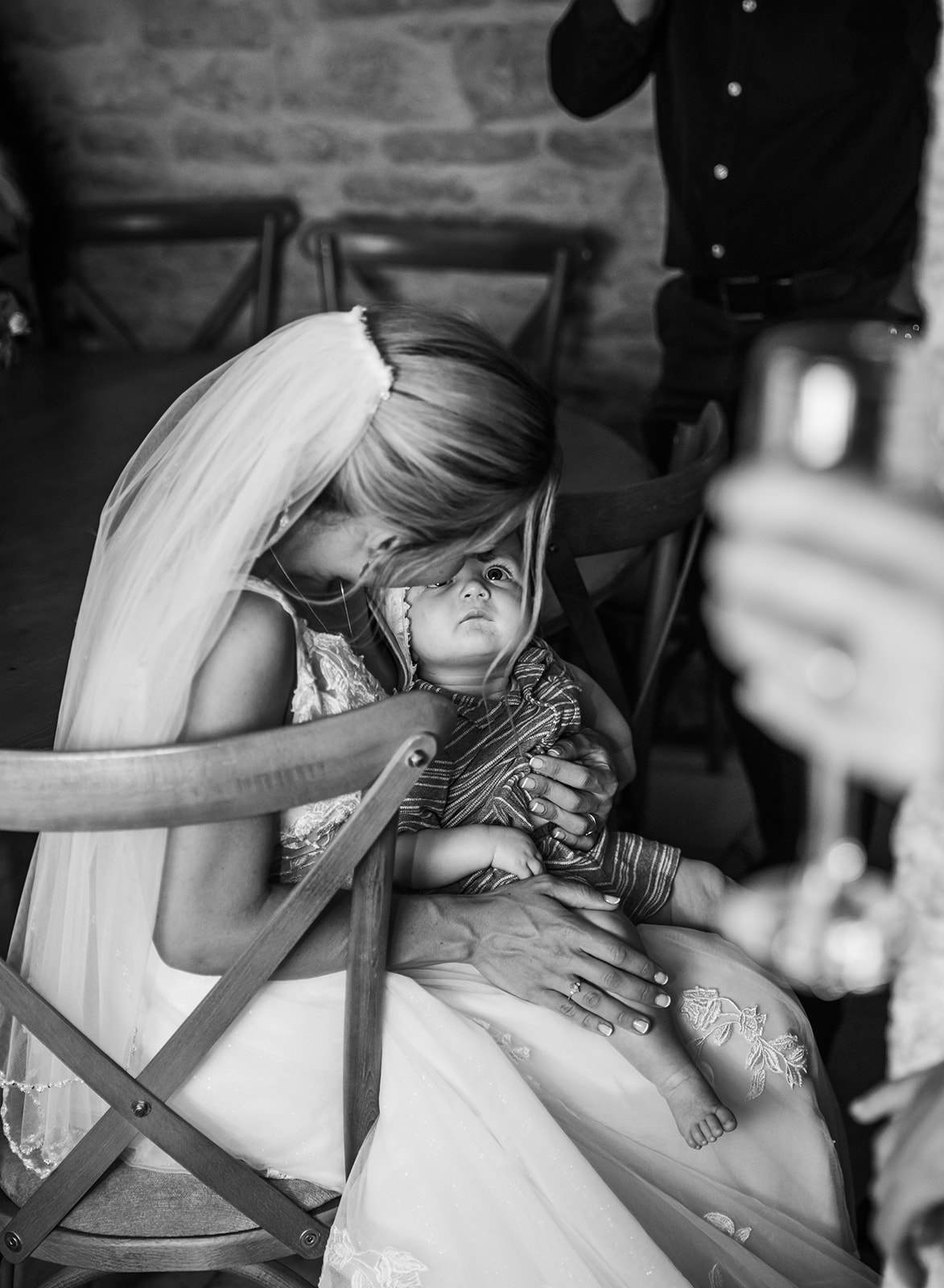 Black and white documentary wedding photography at Kingscote Barn Tetbury, showing a bride sitting and cradling a young child during a quiet moment in the rustic stone barn.