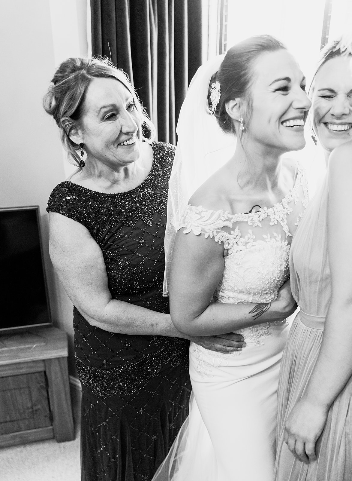 Candid black and white wedding photography in Gloucester, showing a bride laughing with her mother and bridesmaid during morning preparations.