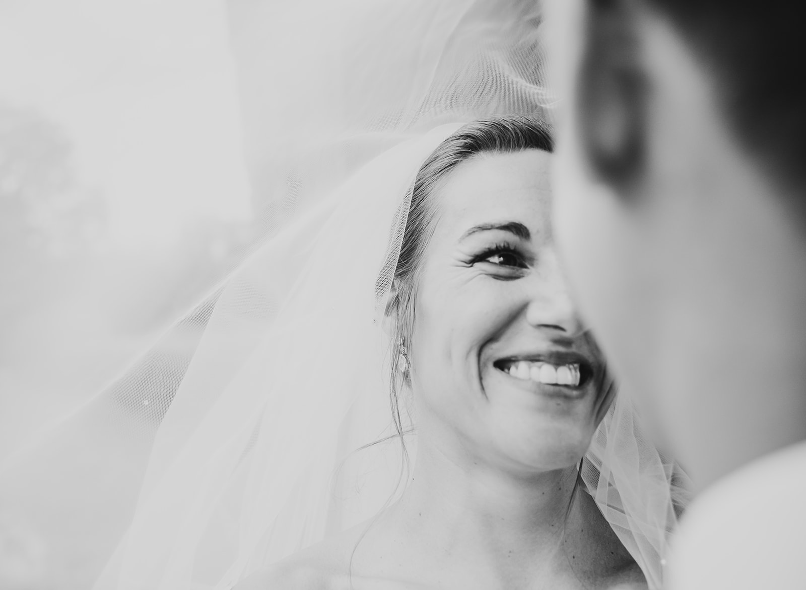 A close-up, high-contrast black and white photograph of a bride smiling radiantly at her groom, framed by the soft texture of her flowing wedding veil.