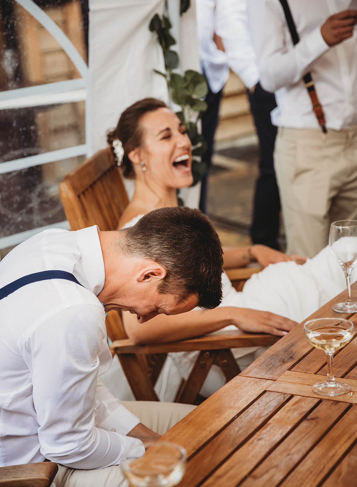 Candid wedding reception photography in Gloucester, capturing a bride and groom in a fit of laughter while seated at a wooden table in a marquee.
