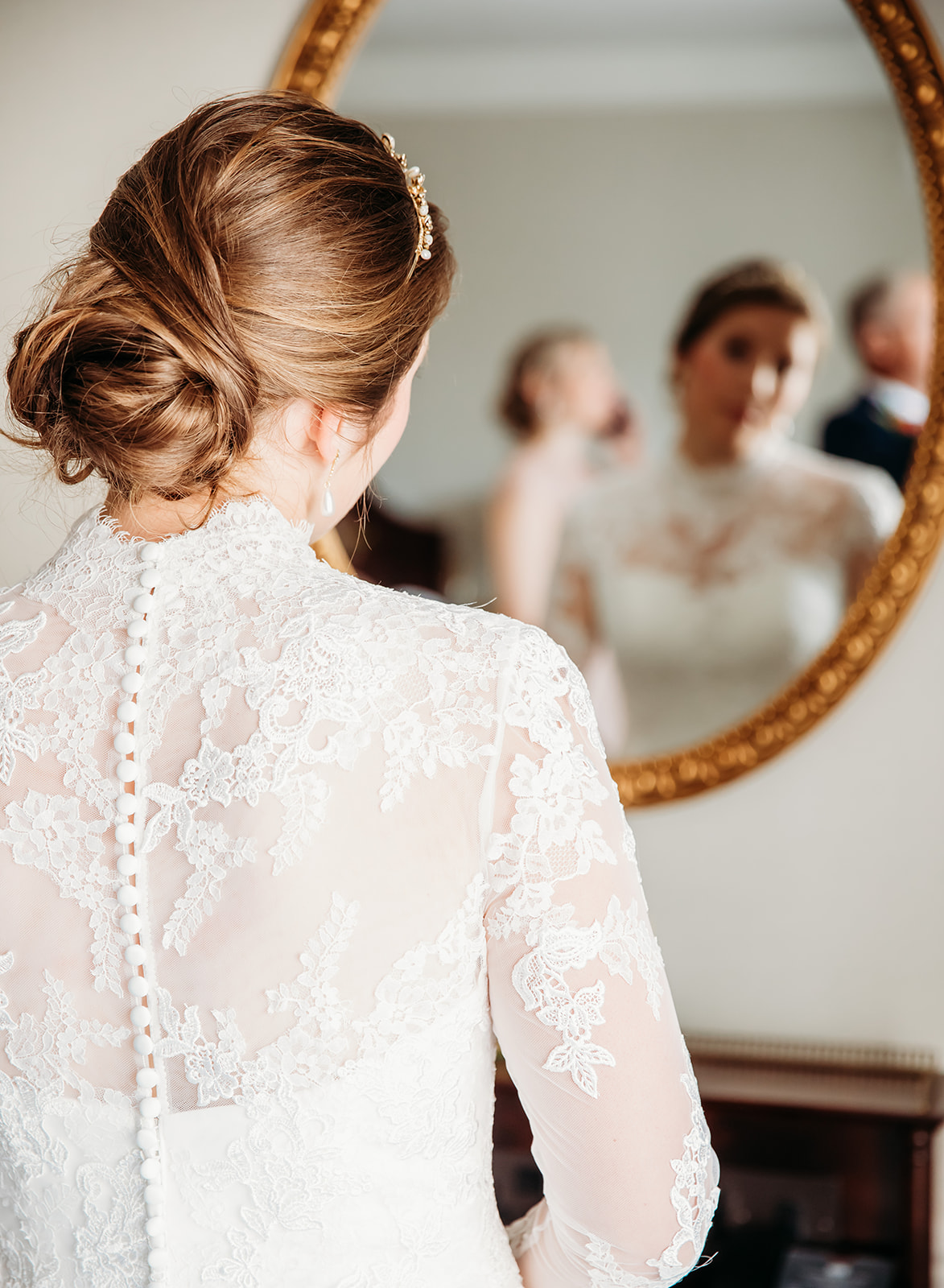 Back view of a bride with an elegant updo and lace wedding dress with button details, looking into an ornate gold mirror at Ellenborough Park.