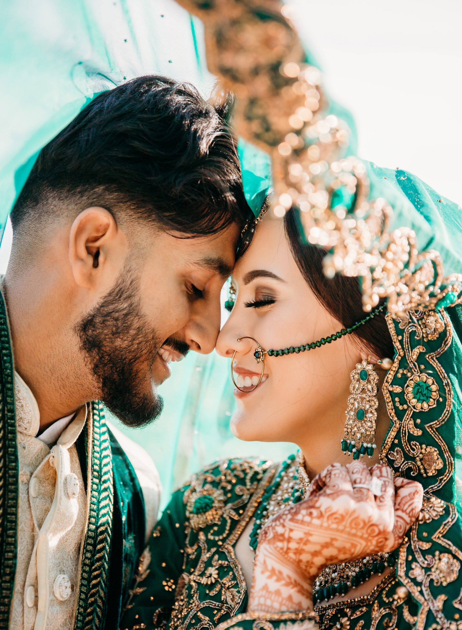 A candid, colorful wedding portrait of a bride and groom in Gloucester smiling together under a sheer green veil, showcasing vibrant traditional attire and jewelry.