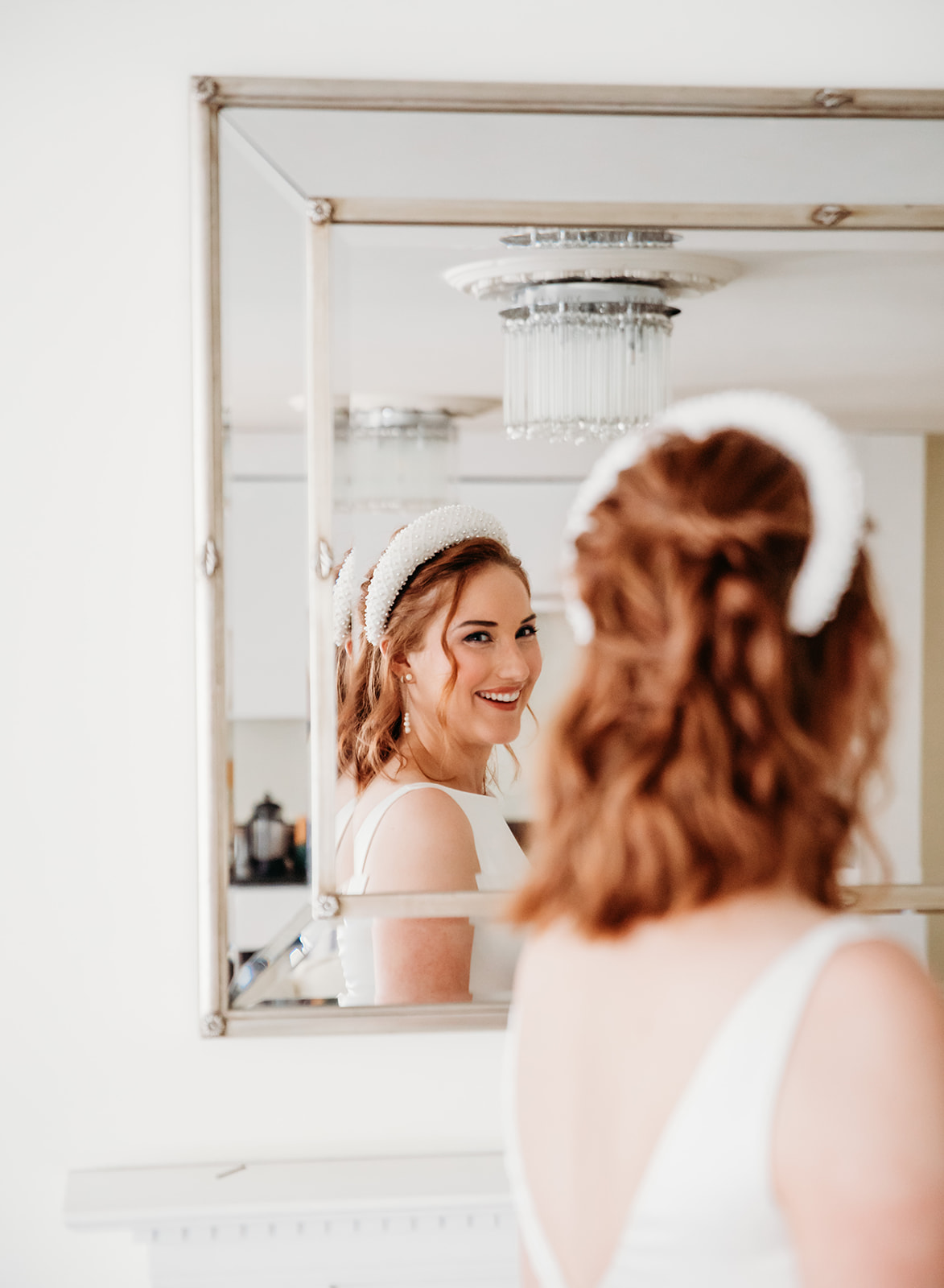 A bride wearing a modern white headband looks at her reflection in an ornate mirror during wedding preparations in Cheltenham, Gloucestershire.