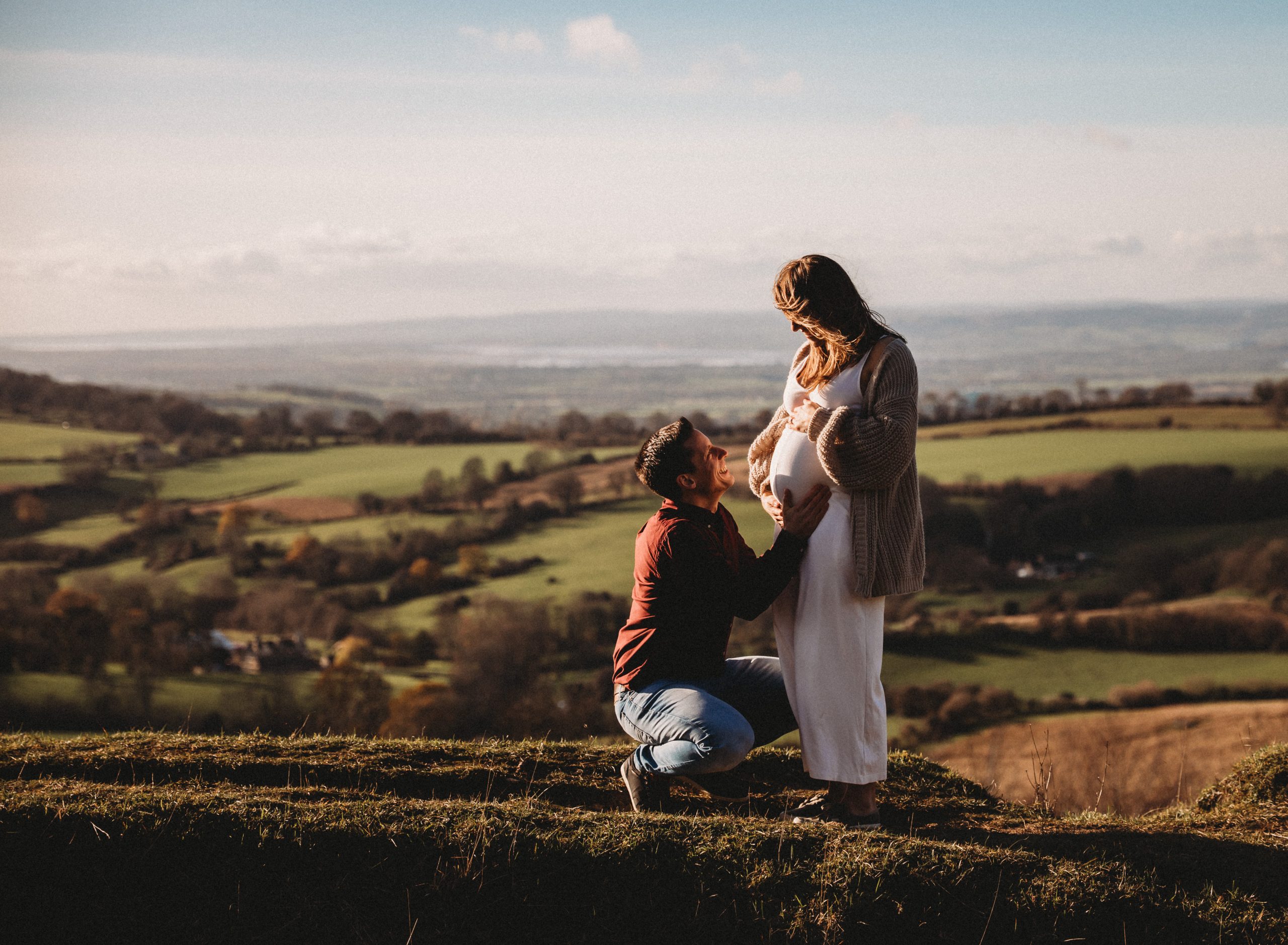 Wide-angle maternity photography of a man kneeling and touching his pregnant partner's belly on a grassy ridge, overlooking a vast, scenic countryside valley at golden hour.