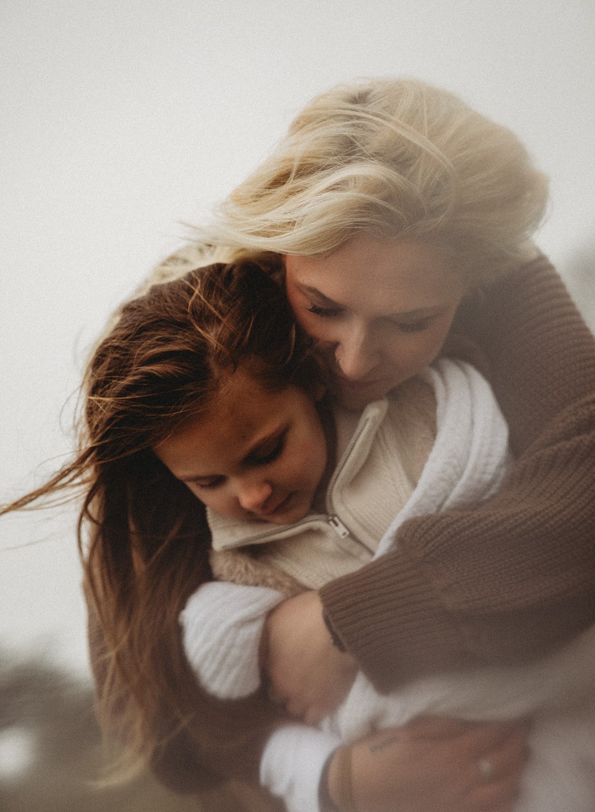 An emotive, grainy lifestyle photograph of a mother and daughter huddled together in a deep embrace, focusing on the intimate bond and natural outdoor lighting.