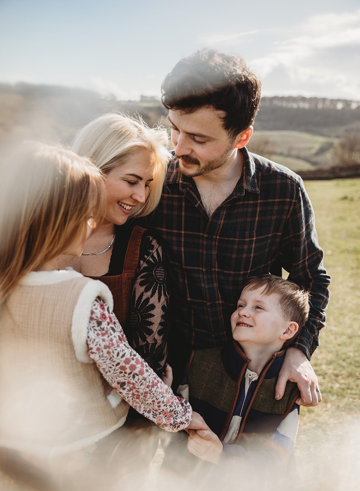 Warm, sun-drenched outdoor family portrait of a mother, father, and two children standing together in a rural meadow.