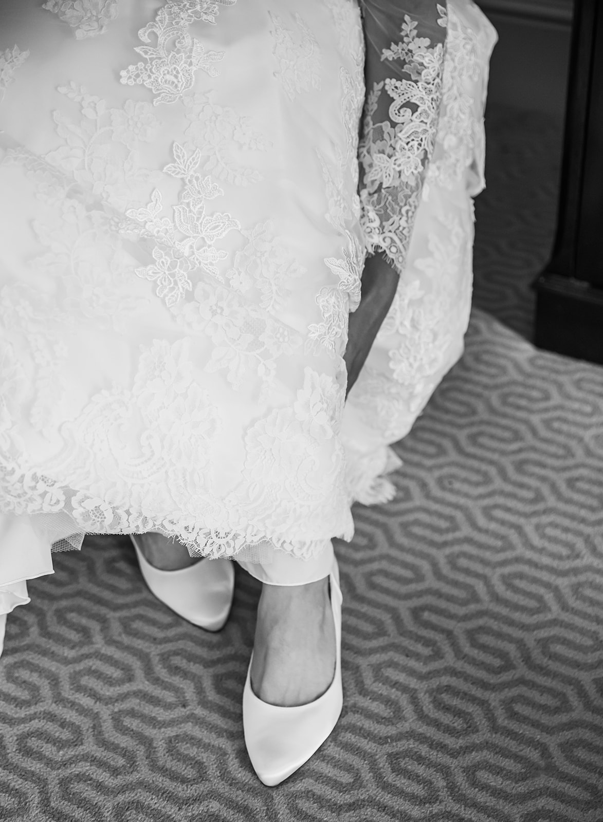 A black and white detail shot of a bride's lace wedding dress hem and silk pointed-toe heels on a patterned carpet at Ellenborough Park Cheltenham.