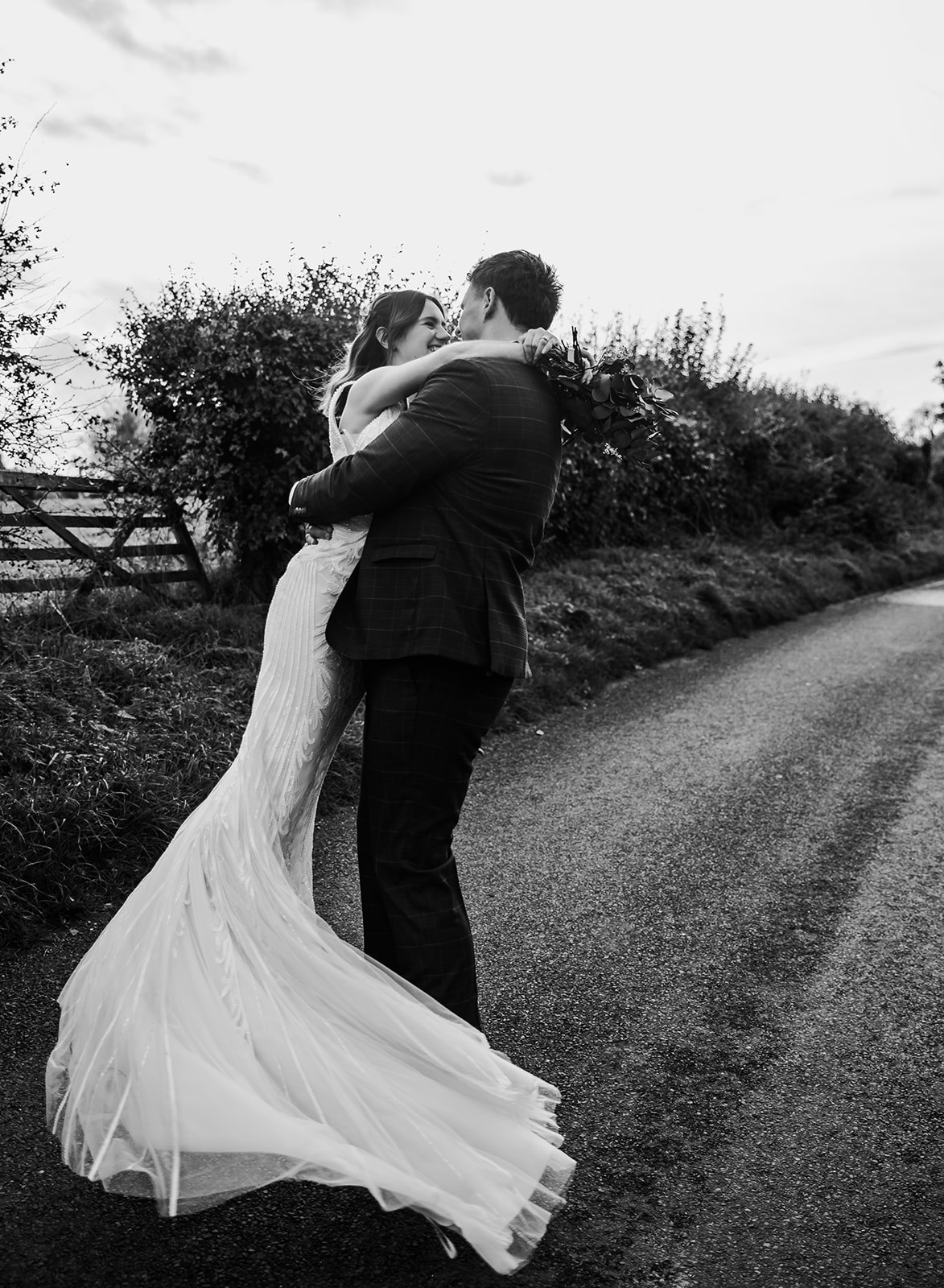 Black and white portrait of a couple embracing on a rural lane at Curradine Barns Worcester, capturing a romantic, film-inspired aesthetic.